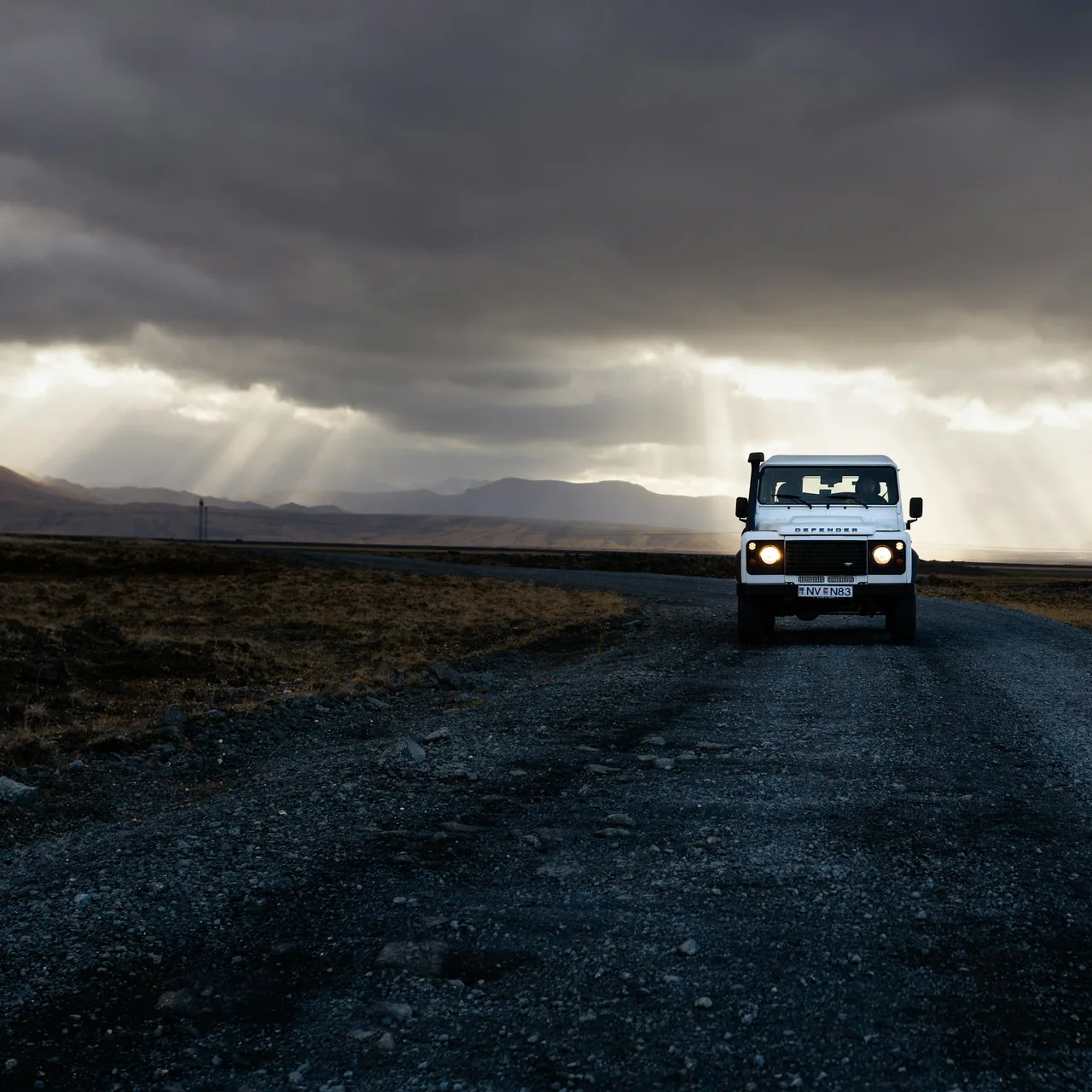 A white Land Rover Defender 4×4 on a remote Icelandic gravel road, with sunbeams breaking through storm clouds over the mountains, photographed by Jon Flobrant
